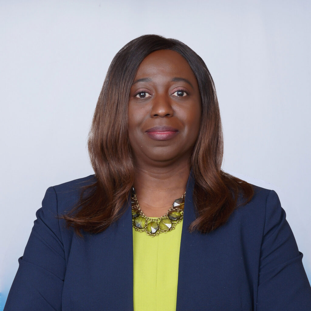 A woman with straight, shoulder-length brown hair wearing a navy blazer over a bright yellow top and a statement necklace, poses in front of a plain light background, looking directly at the camera.