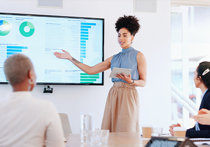 A woman stands and presents data on charts displayed on a large screen to three seated colleagues in a bright meeting room, holding a tablet and gesturing toward the screen.