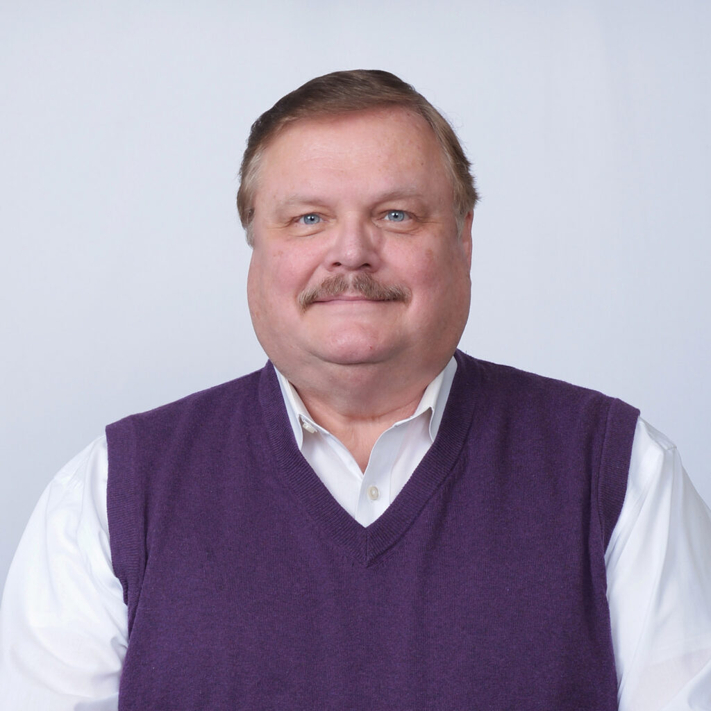 A middle-aged man with short light brown hair and a mustache, wearing a white dress shirt and a purple sweater vest, poses in front of a plain light background.