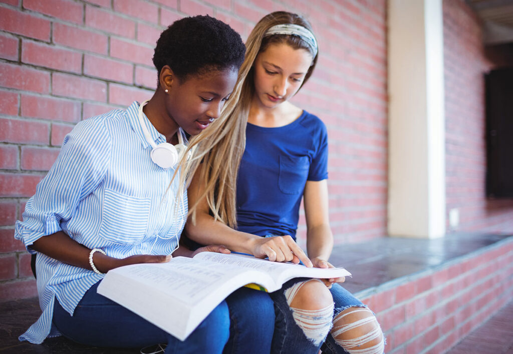 Two teenage girls sit together by a brick wall, looking at an open book. One girl points at the pages while the other reads, both appearing focused and engaged in studying.