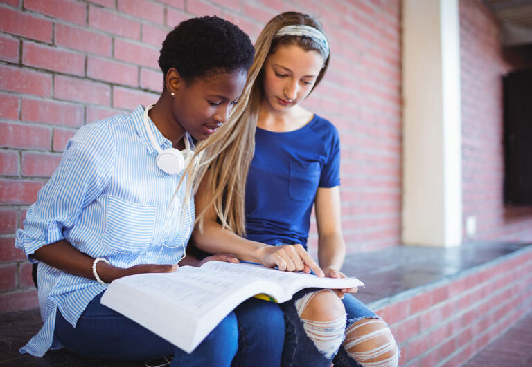Two teenage girls sit together by a brick wall, looking at an open book. One girl points at the pages while the other reads, both appearing focused and engaged in studying.