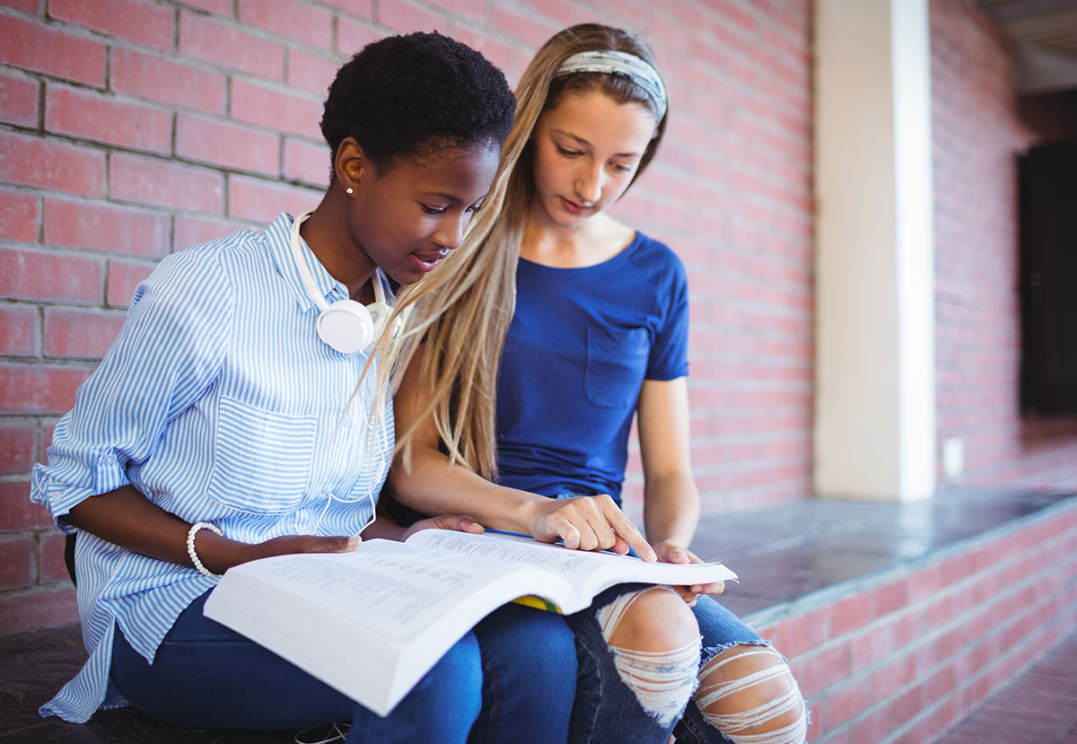 Two teenage girls sit together by a brick wall, looking at an open book. One girl points at the pages while the other reads, both appearing focused and engaged in studying.