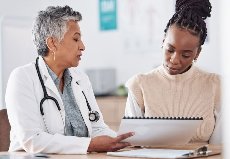 A doctor with gray hair and a stethoscope talks to a patient, showing her information on a clipboard. The patient, wearing a beige sweater, looks down attentively at the papers in a medical office.