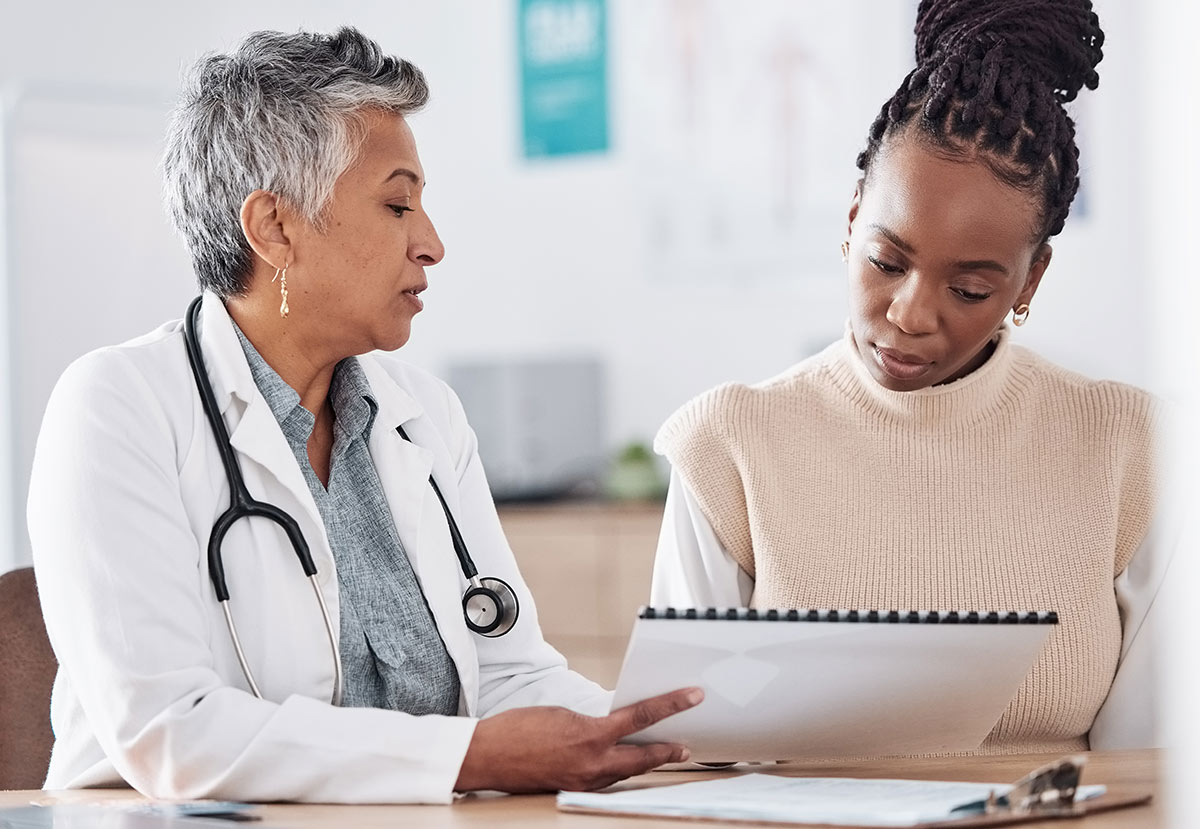 A doctor with gray hair and a stethoscope talks to a patient, showing her information on a clipboard. The patient, wearing a beige sweater, looks down attentively at the papers in a medical office.
