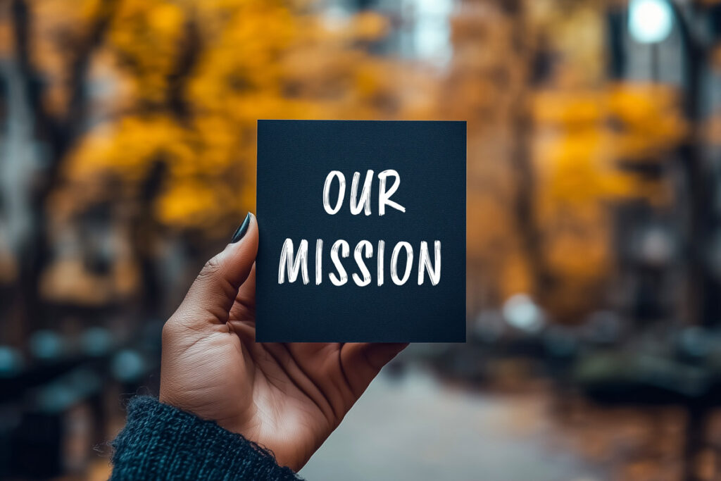 A hand holds a black card with the words OUR MISSION written in white, with a blurred outdoor background featuring autumn trees and fallen leaves.