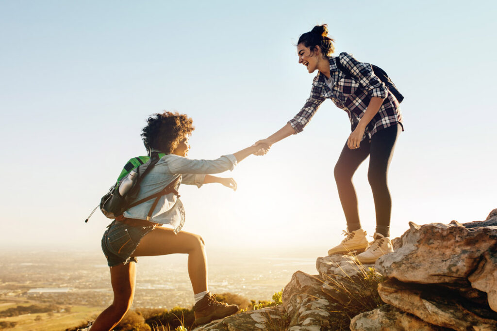 Two women hiking on a rocky hill, one reaching down to help the other up. They both wear backpacks and casual outdoor clothing, with a scenic landscape and clear sky in the background.