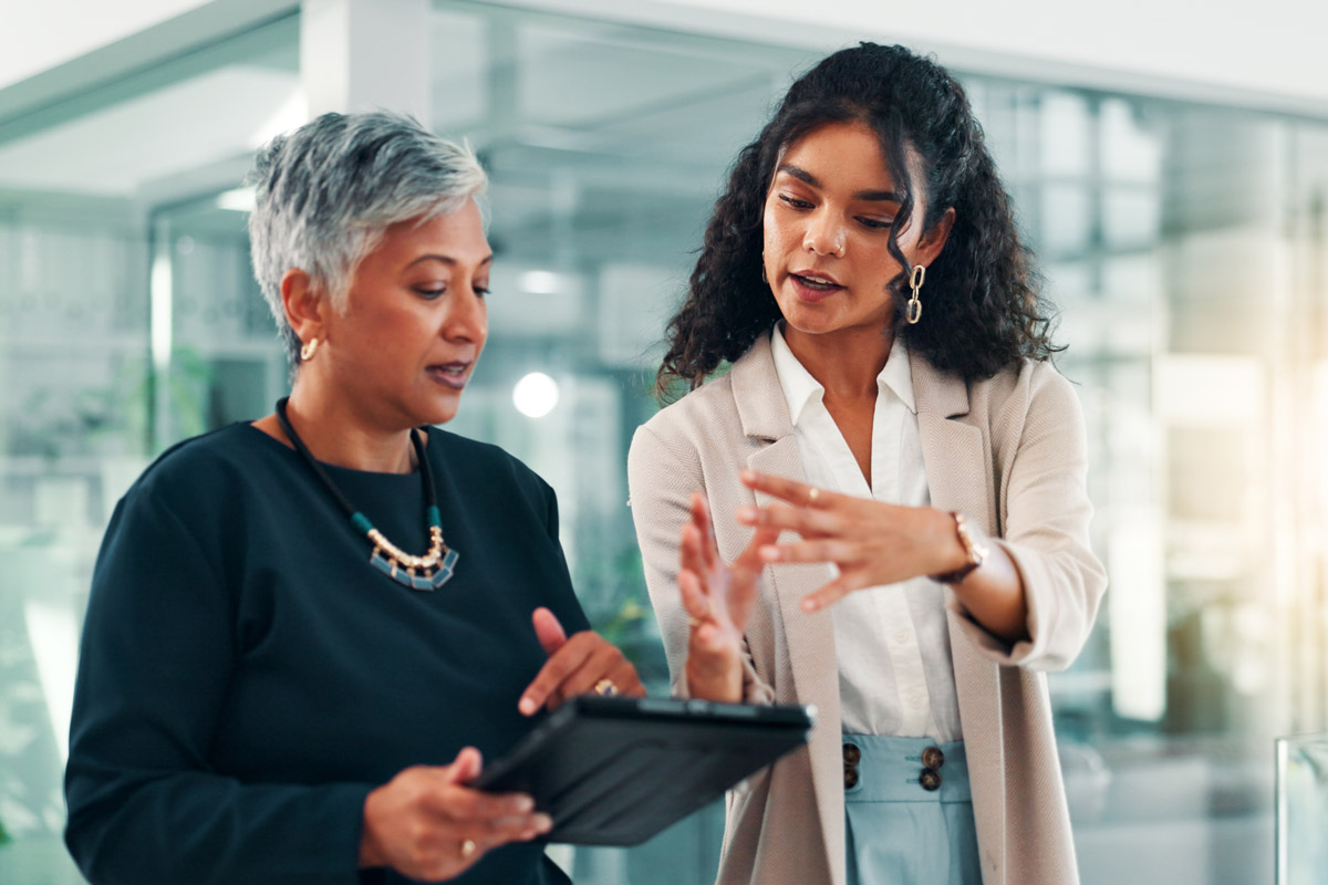 Two women in business attire stand in an office; one is holding a tablet while the other gestures with her hands as they discuss something, appearing focused and engaged in conversation.
